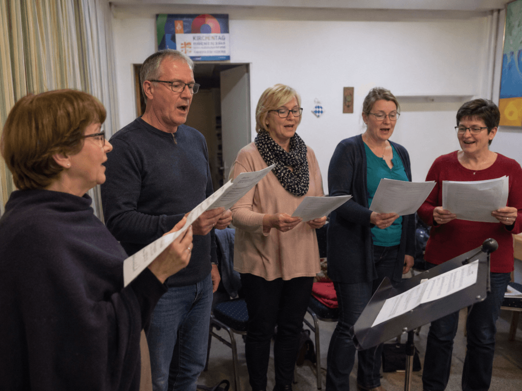 Erwachsene singen gemeinsam in einer kleinen Chorprobe und halten Notenblätter in den Händen