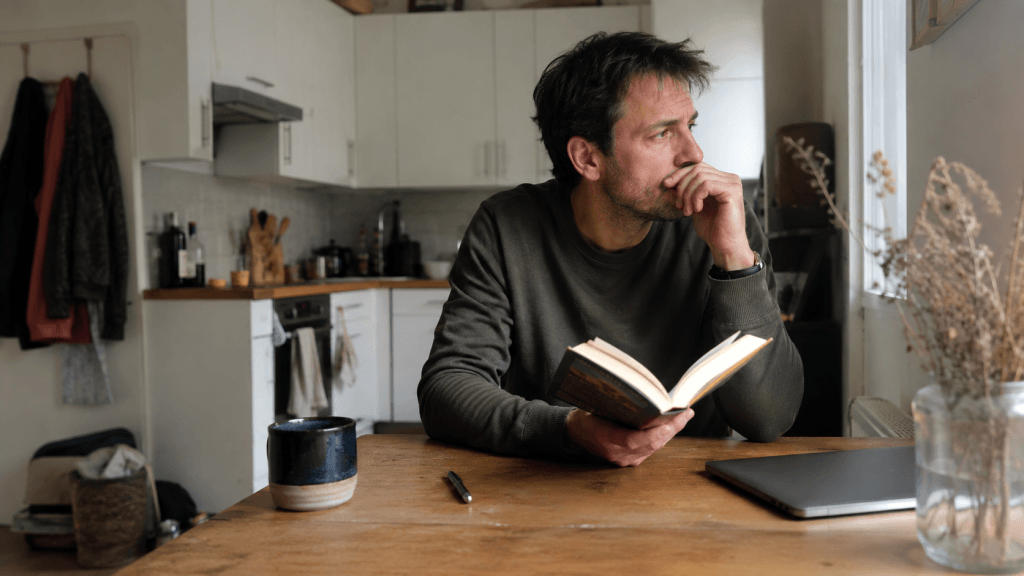 Nachdenklicher Mann mit Buch am Küchentisch als Beispiel für eine Lesegewohnheit, die im Alltag schwer umzusetzen ist.